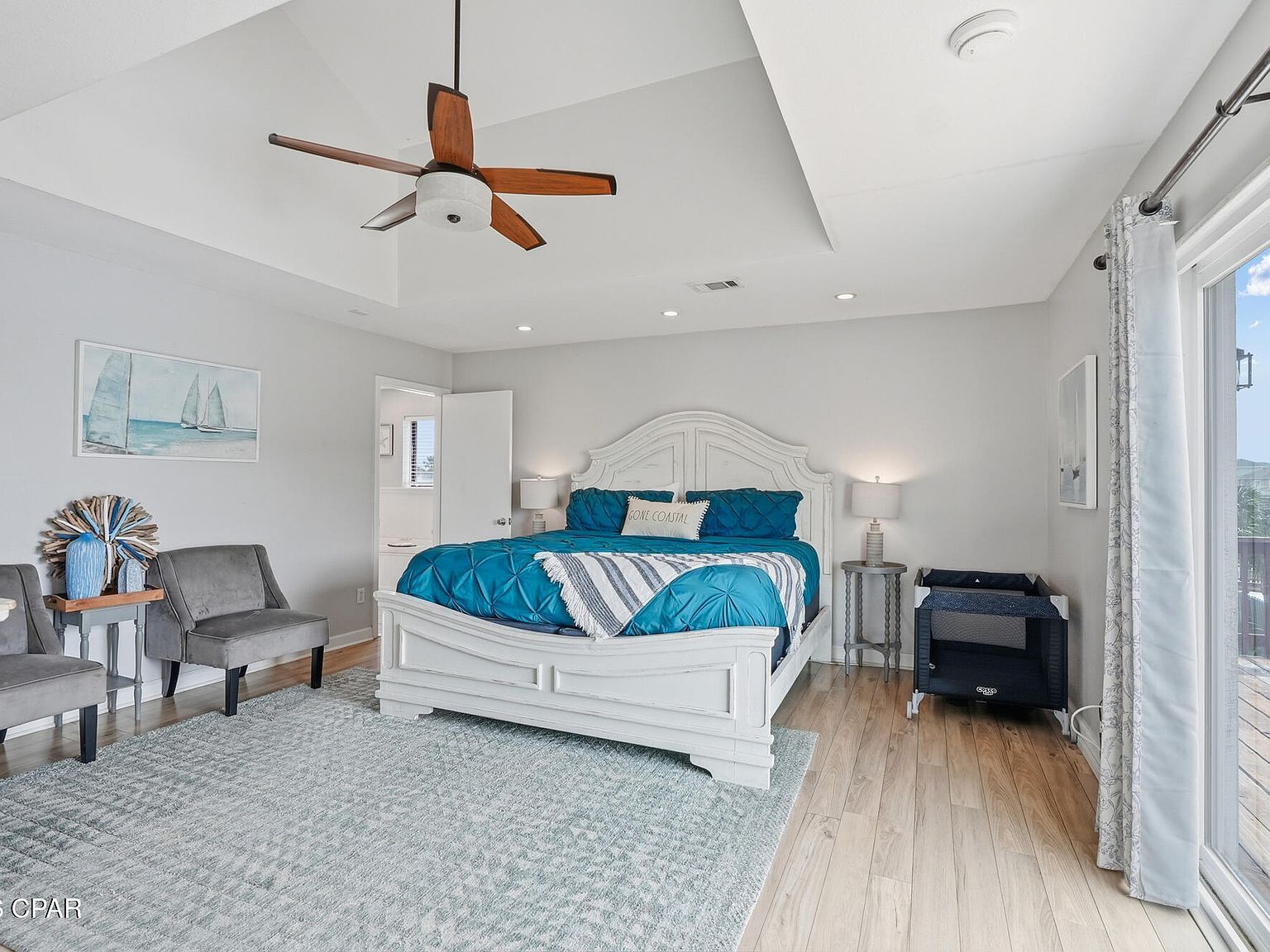 A guest bedroom with a white bed, teal bedding, sailboat art, light wood floors, and a door to the deck and palms