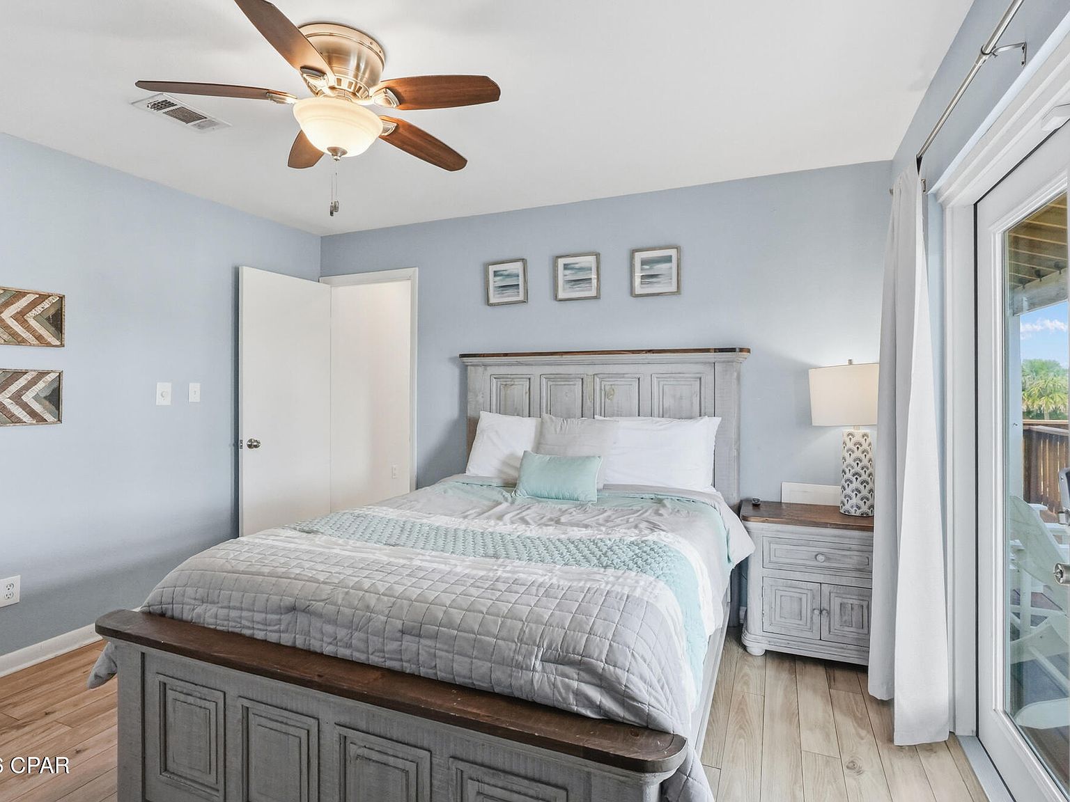 A powder-blue guest bedroom with a rustic gray bed, a wood dresser, a ceiling fan, and a slider to the deck