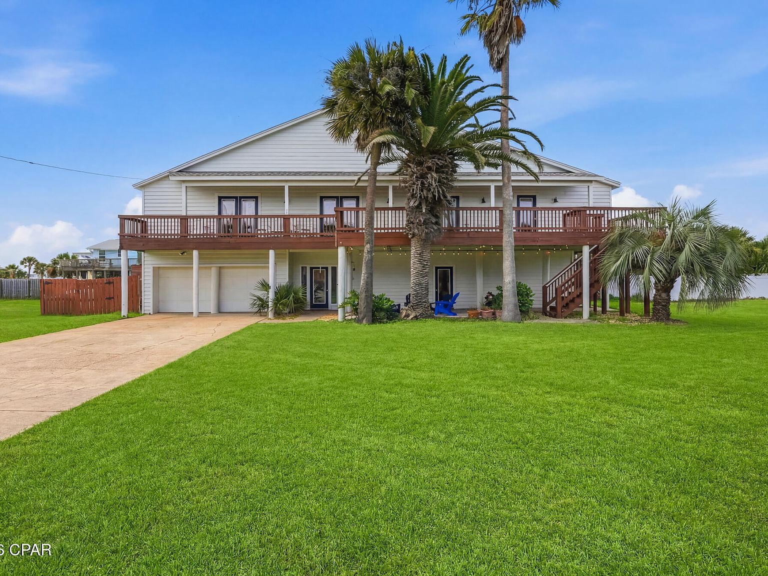 Front elevation of 1205 Via De Luna Dr, a two-story coastal beach home with a wraparound deck, white siding, and date palms over a green lawn in Pensacola Beach